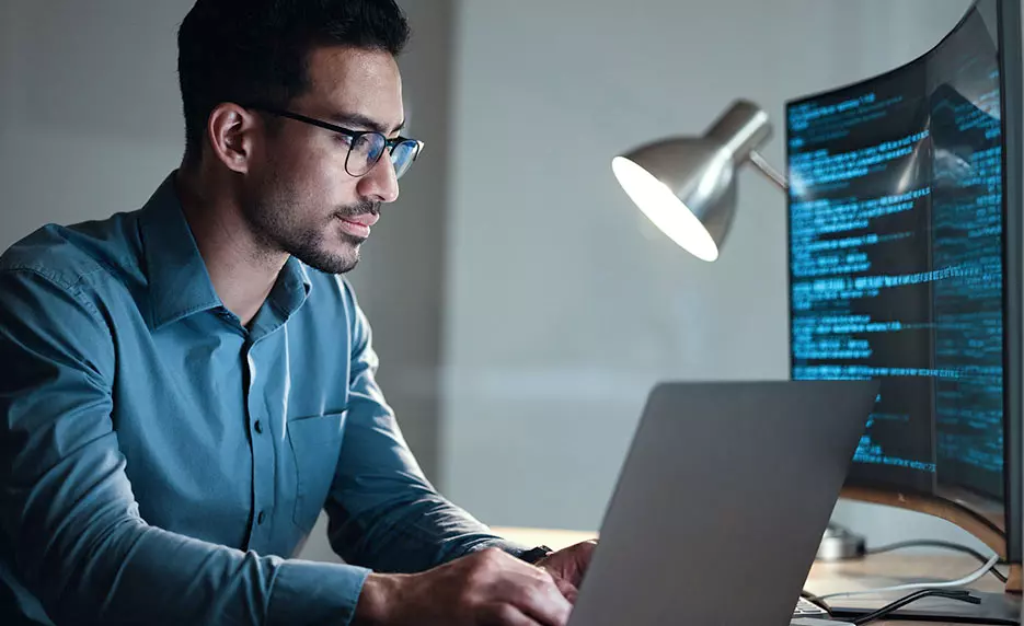 man working at a desk with a desktop computer showing code and a laptop